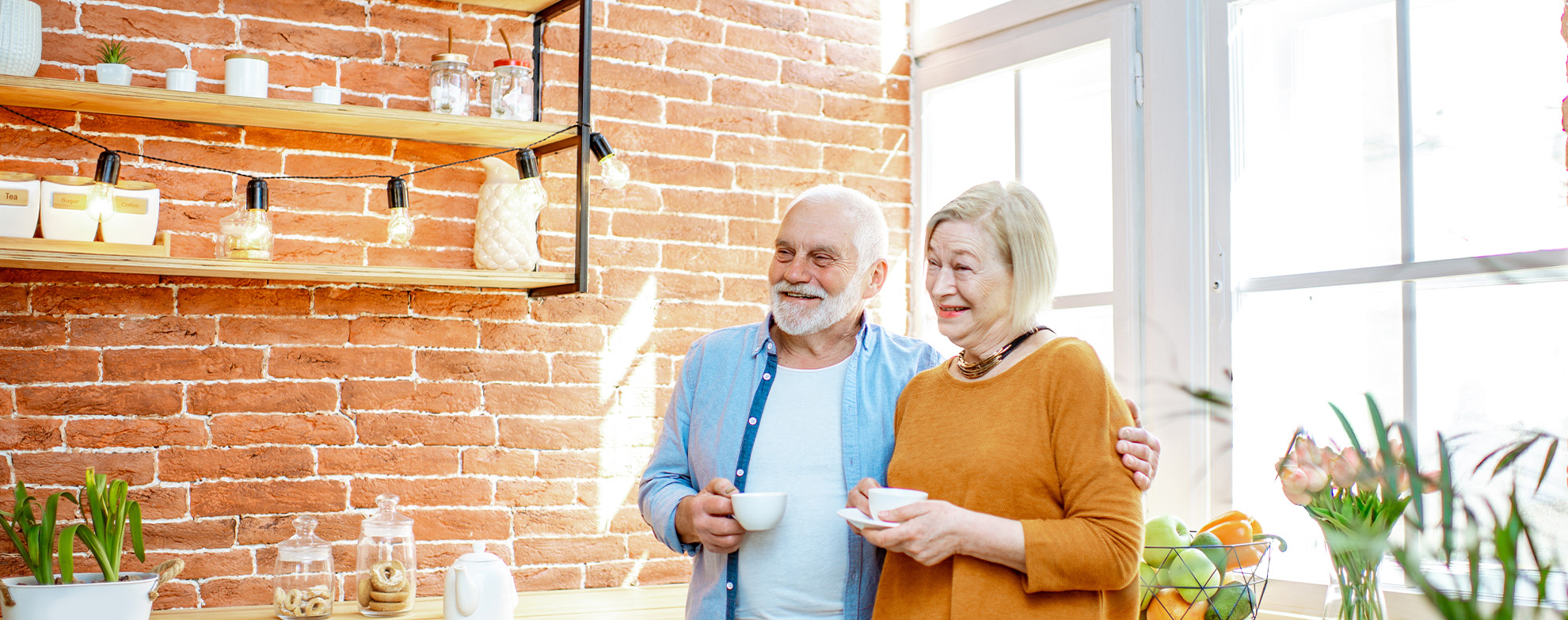 Senior couple standing together with coffee cups near the window on the kitchen at home